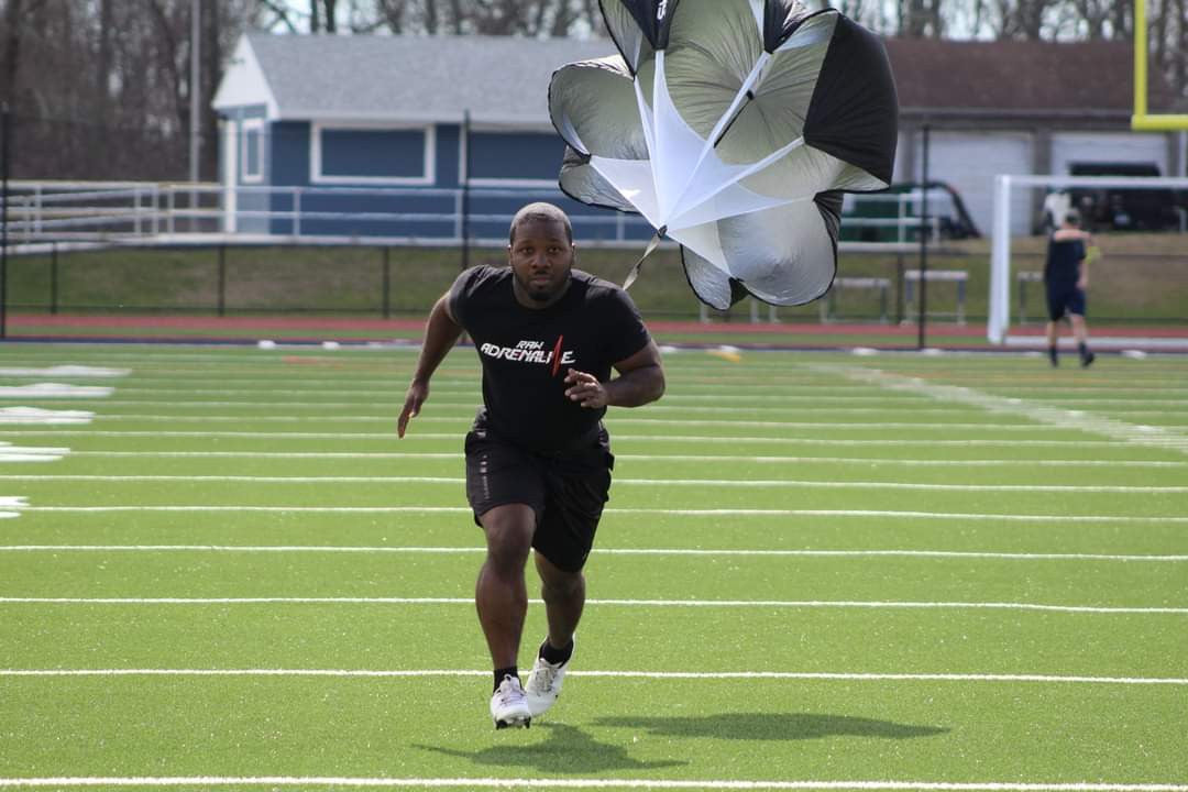 Runner in a black Raw Adrenaline T-shirt sprinting across a field with a resistance parachute.