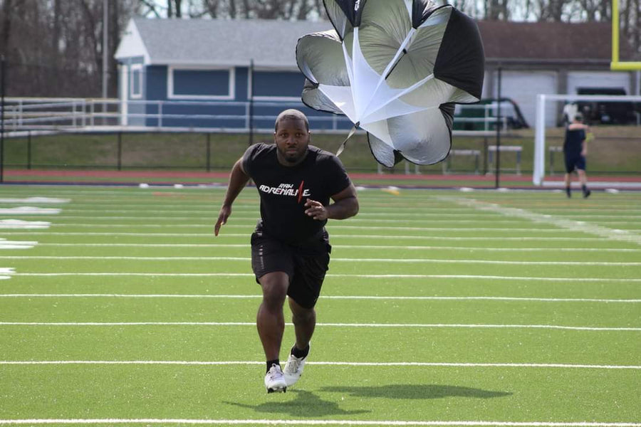 Runner in a black Raw Adrenaline T-shirt sprinting across a field with a resistance parachute.