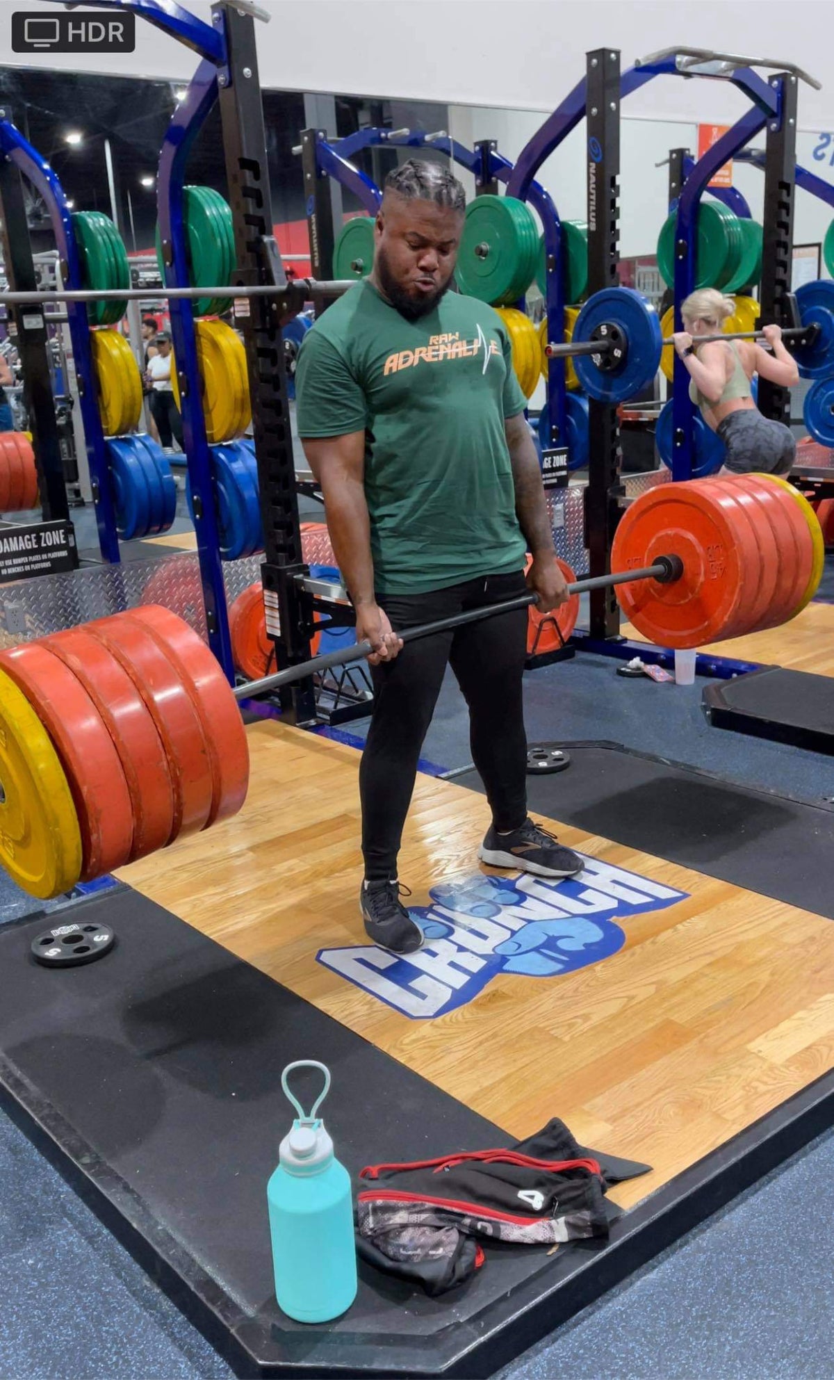 Lifter wearing a green Raw Adrenaline T-shirt and holding a barbell in a weight room.