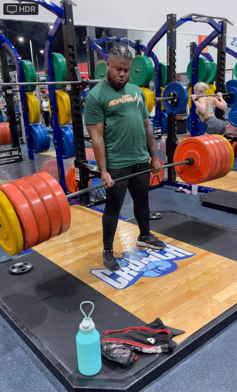 Lifter wearing a green Raw Adrenaline T-shirt and holding a loaded barbell in a weight room.